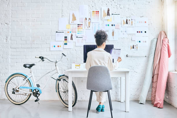 istock Designer woman, workshop and desk by wall, moodboard and reading documents with sketch, planning or idea. Startup fashion expert, studio and sitting at table for small business growth, vision or goal 1496871692