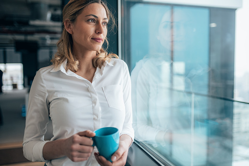Thoughtful businesswoman looking through the window. Beautiful business woman standing in the office and holding cup of coffee.