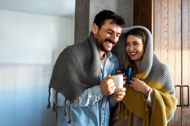 istock Happy young couple with hot drinks cuddling under warm blanket at home 1496877123