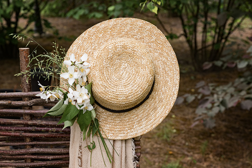 A beautiful straw hat with a bouquet of white flowers hangs on the fence. Nature and garden summer background. Nobody