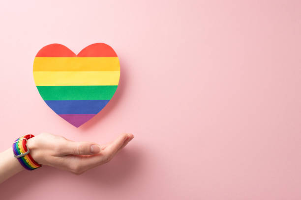 foto della mano femminile che indossa un braccialetto simbolico e rilascia un cuore arcobaleno su uno sfondo rosa pastello, perfetto per un concetto di parata lgbt - gay pride wristband rainbow lgbt foto e immagini stock
