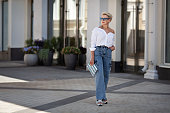 Rejuvenated Trendy mature woman with short hair stands outdoors in city street wears youth clothing white shirt, jeans cargo pants, clutch. Urban style fashion, age and timeless beauty