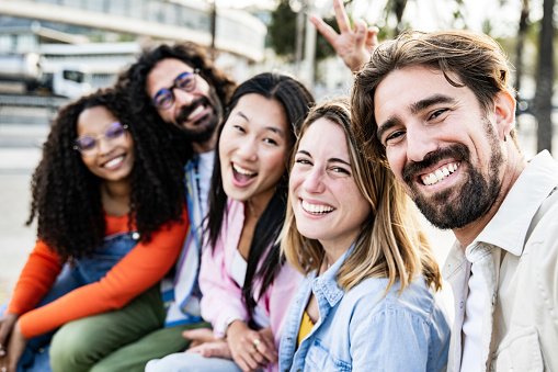 Diverse group of friends taking a selfie in the street sitting and looking at camera. Cheerful multiracial group of young hipsters taking a picture outside. Focus on front man and woman.