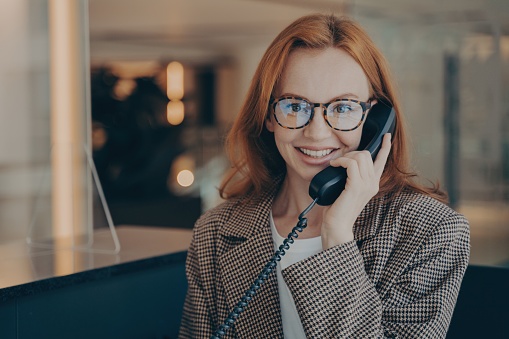 Smiling redhead office worker in plaid jacket and glasses, discussing project completion date on landline phone, office background blurred.