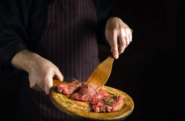 istock The chef turns over raw beef steaks with a wooden spatula before barbecue. Preparation for roasting meat with rosemary and spices. Black space for hotel recipe or menu. 1496899341