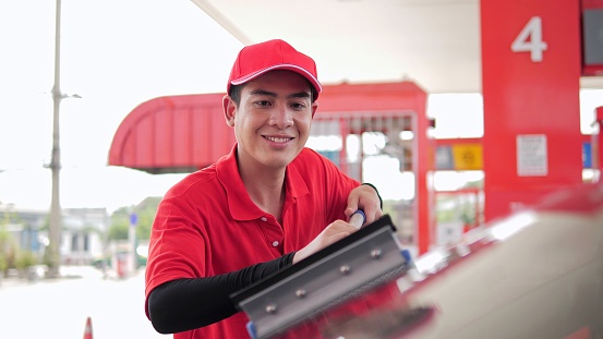 Young asian male gas station worker provides windshield cleaning. Cleaning the windshield of a consumer car while filling up at gas station. Transportation power business concept