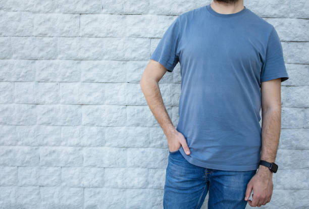 Man in a blue blank T-shirt, jeans, a gray wall. Close-up, no face. The background is a brick plain wall. Mockup, empty space. Front view. Adult male 30-35 years old. Horizontal layout