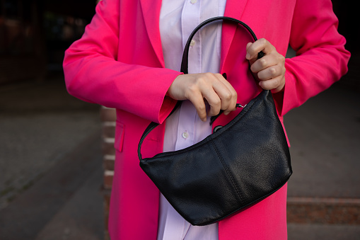 Woman no face. Looking for her phone or handbag in her trendy fashionable black leather bag. Pink blazer, a lilac shirt on the street. Street style, outfit. Opens his bag. Bag in the style of the 90s