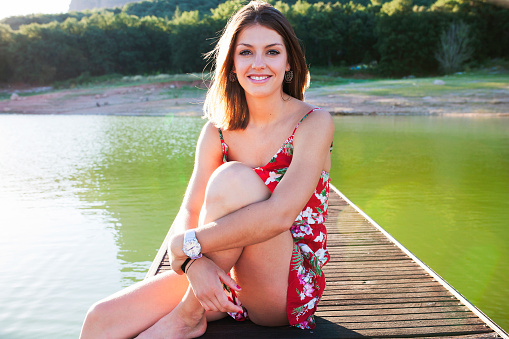 Young happy smiling brunette woman in dress sitting on wooden pier posing and looking at camera in sunlight.