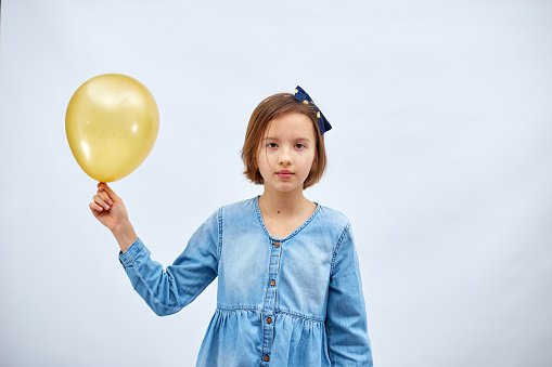 Pretty sad little girl in casual denim dress hold in hand yellow balloon on white background, studio