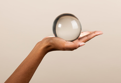 Unrecognizable African American female demonstrating transparent glass sphere resembling magic crystal ball against beige background
