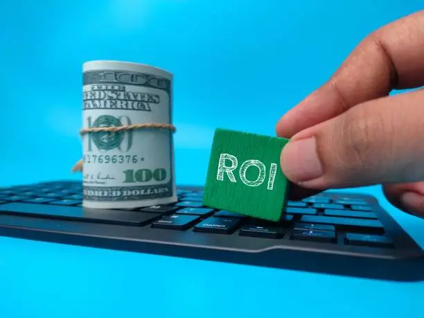 A mature adult man with a green rubber block with the ROI printed on near a roll of dollars resting on a computer keyboard
