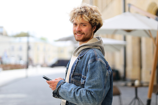 A young guy in casual clothes holds a mobile phone in his hands and smiles against blurred street view.