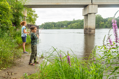 A young brother and sister fishing near a bridge while it is raining.