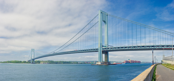 High resolution stitched panorama of the Verrazano-Narrows Bridge on a Cloudy Summer Morning with Promenade and Container Cargo Ship. The bridge connects boroughs of Brooklyn and Staten Island in New York City. The bridge was built in 1964 and is the largest suspension bridge in the USA. Historic Fort Wadsworth is under the bridge. The photo was taken from the Shore promenade in Bay Ridge Brooklyn. Canon EOS 6D full frame censor camera. Canon EF 50mm F/1.8 II lens. 2.13:1 Image Aspect Ratio. This image was downsized to 50MP. Original image resolution is 64MP or 11668 x 5466 px.