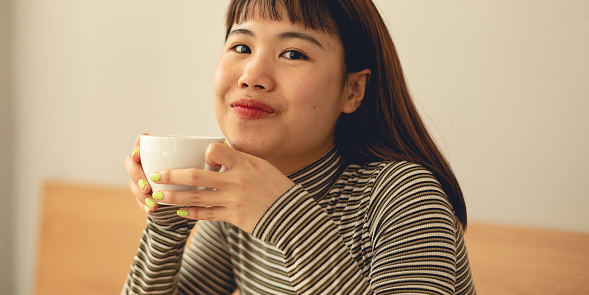 Female portrait of asian woman indoors with drink in cafe. Photo of girl with beverage. Person is drinking coffee from white cup. Lifestyle photography with people.