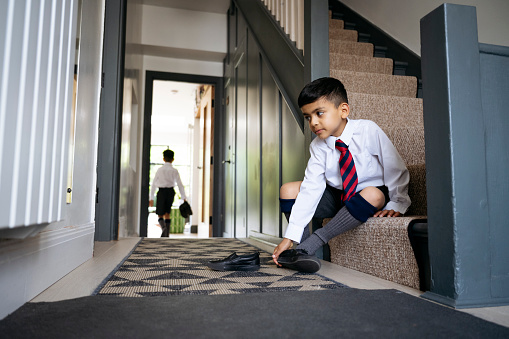 Full length view of 5 year old boy in uniform sitting at bottom of staircase in family home and putting on shoe, 7 year old boy in background.