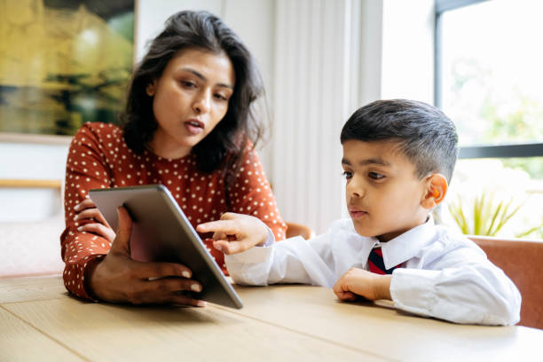 Mother assisting schoolboy with homework on portable device Waist-up view of 5 year old student in uniform sitting with early 40s woman at dining table in family home and talking as they use digital tablet. english spoken stock pictures, royalty-free photos & images