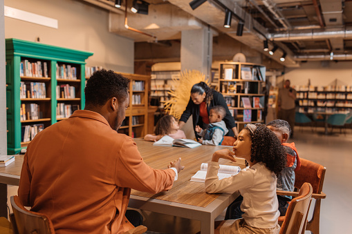 A black teacher reading a book to young black and diverse school children in a library