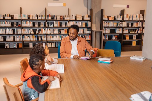 A black teacher reading a book to young black and diverse school children in a library