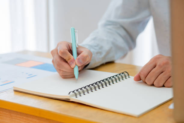 istock Closeup worker planning with pen, notes and schedule of information in journal, reports or logistics agenda, strategy and online data analysis at desk. Worker, computer and notebook 1496939564