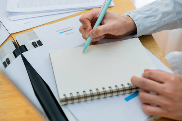 istock Closeup worker planning with pen, notes and schedule of information in journal, reports or logistics agenda, strategy and online data analysis at desk. Worker, computer and notebook 1496939628