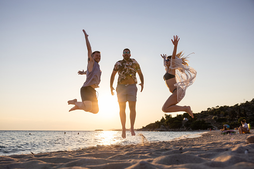 Happy and playful young Caucasian female and male friends, jumping at the beach during sunset on their vacation