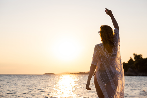 Rear-view of an unrecognizable young Caucasian woman in swimwear and sundress, dancing on the beach while admiring the sunset above the sea