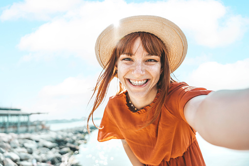 Smiling young woman taking a selfie at beach looking at camera - Cheerful caucasian tourist enjoying at the sea during holiday in a sunny day - Travel Happiness concept