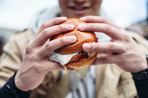 Focus on foreground as Gen Z Londoner smiles before biting into pork and egg street food roll from market stall.