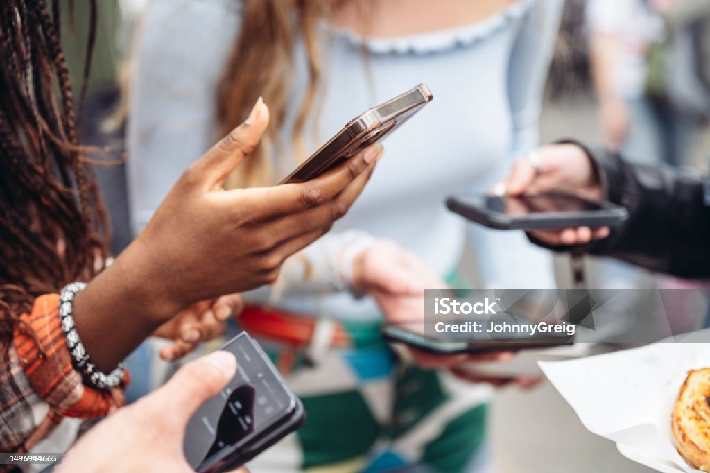 Gen Z Londoners holding smart phones using social media Focus on teenage Black woman standing with friends, all using their portable devices. Asian and Indian Ethnicities Stock Photo
