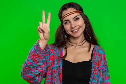 Young woman showing victory sign, hoping for success and win, doing peace gesture, smiling with kind optimistic expression. Pretty hippie girl isolated alone on chroma key background, green screen