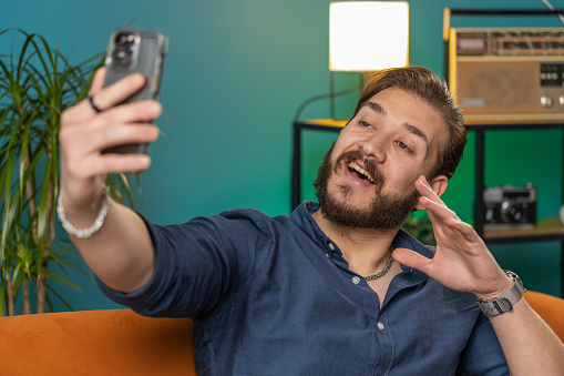 Happy lebanese man blogger taking selfie on smartphone, communicating video call online with social media subscribers followers, recording vlog stories. Young arabian guy at home apartment on couch