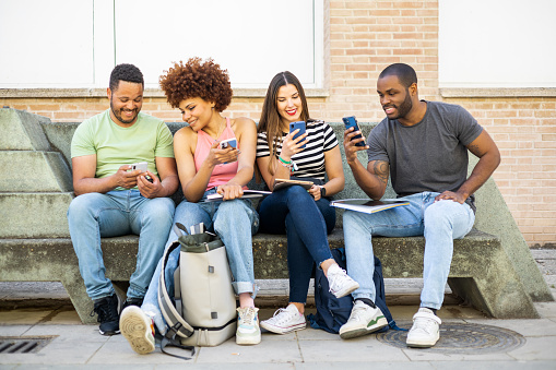 A group of multi-ethnic college students are sitting on a bench outside the university each interacting with their mobile. Concept of disconnecting from class, wasting time with social networks.
