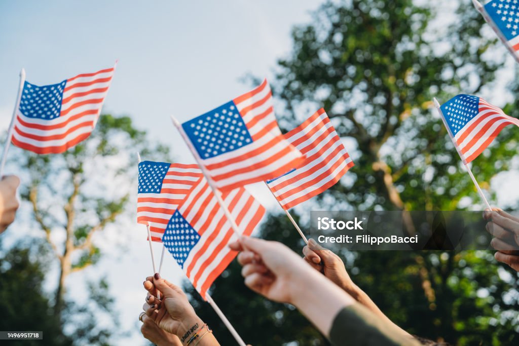 A group of people is waving small American flags at sunset A group of people is waving small American flags at sunset. Concept for various topics like Happy Veterans Day, Labour Day, Independence Day. American Flag Stock Photo