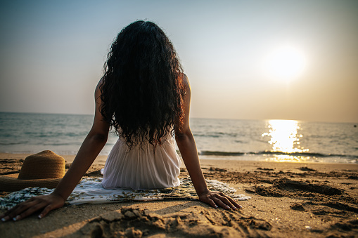 Rare view Beautiful young woman with long curly hair in white dress sitting on the sand, She looked sunset in the sea while relaxing on her summer vacation. Copy space to insert your advertisement.
