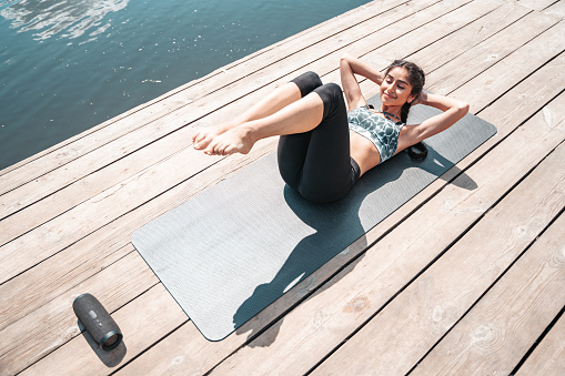 Young Indian woman doing abs exercise on wooden pier by the river enjoying summer morning fresh air