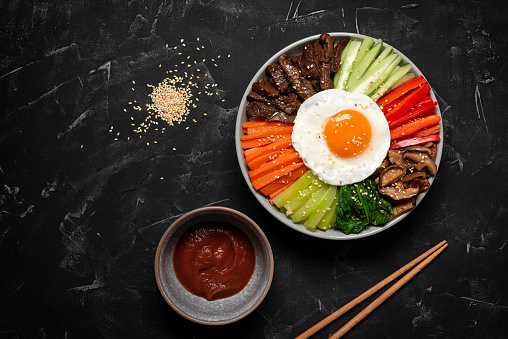 Korean Bibimbap on a black stone background. Rice with beef, vegetables, mushrooms and fried egg in a bowl. Top view, flat lay.
