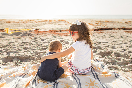 A 1-Year-Old Cuban American Baby Boy Wearing a Navy Blue Cotton Romper Sitting with His 4-Year-Old Sister Wearing a White T-Shirt, Striped Shorts, Sunglasses & a Bow While on a Blue Striped Turkish Towel on a Sandy Beach in Singer Island, Florida at Sunrise in 2023