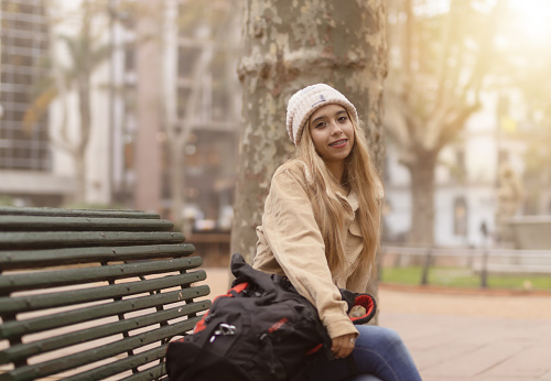 Young woman wearing a hat sitting on a square bench with her travel backpack. Travel alone concept.