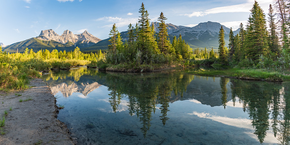 Panoramic mountain peaks reflecting in the calm water below at Three Sisters in Canmore, near Banff National Park, Canada in summer time with wilderness, wild, tourist, tourism area, panorama.