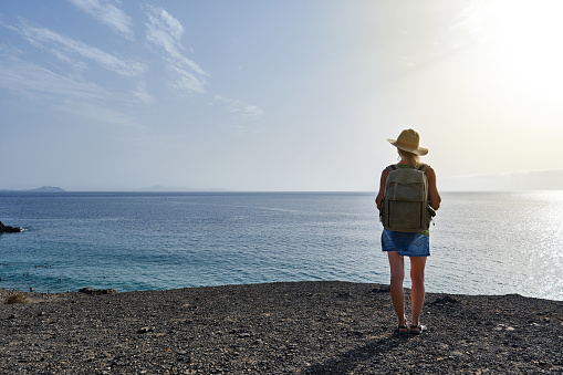 Rear view of a woman with a hat and backpack in front of a cliff overlooking the sea.