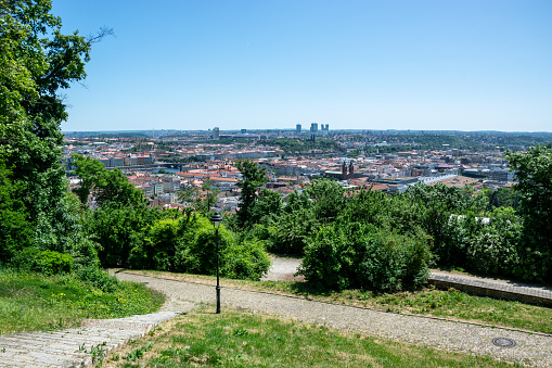 A panoramic view of Prague, Czech Republic, with red rooftops and a blue sky. The photo is taken from a high vantage point, looking down on the city. The buildings are mostly traditional European style, with orange and red rooftops. The sky is blue with some clouds. In the background, you can see the Prague Castle and St. Vitus Cathedral.