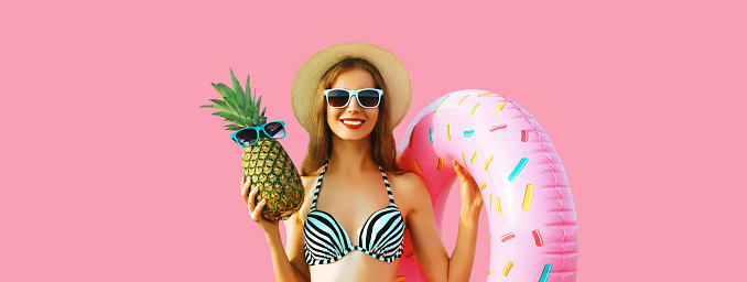 Summer vacation, tourism, happy smiling young woman with swimming inflatable ring and pineapple wearing straw hat on studio pink background