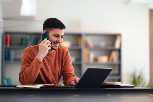 Happy business man speaking on the phone with a client while typing something on a laptop computer keyboard and sitting at his office desk.