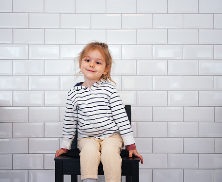 Smiling little girl looking at camera while sitting in front of white tiles wall.