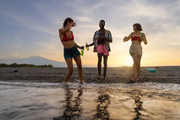 An Afro-American man in colorful shirt and swimwear, and two Caucasian women in bikinis and shorts, dancing, joking, and having fun on the beach at sunset while holding beer, waves touching their feet