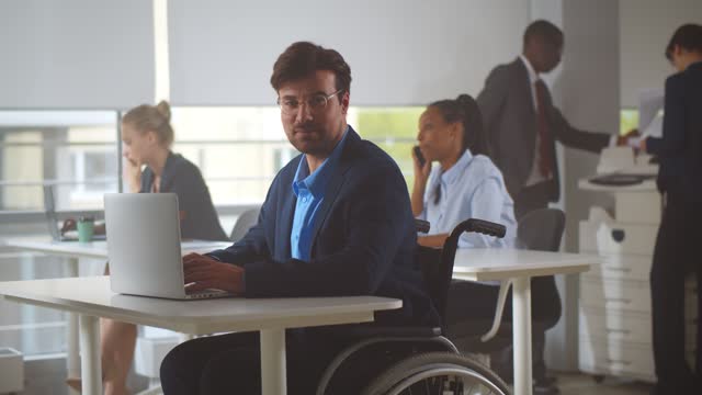 Handicapped office manager looking at camera sitting in modern startup company office in wheelchair