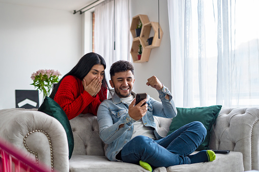 Excited Latin couple watching cell phone to win online lottery prize, gather in the living room of the apartment.