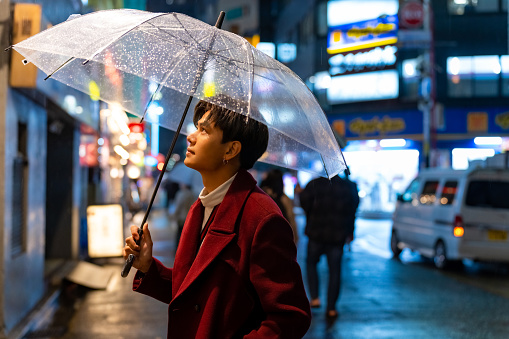 Young Asian man holding umbrella walking city street with night lights and crowd of people in Tokyo city, Japan in raining night. Handsome guy enjoy outdoor lifestyle travel in the city on vacation.